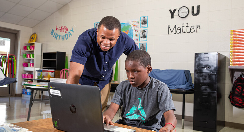 A teacher and student looking at a laptop screen together in a classroom with a large sign behind them that says "You Matter"