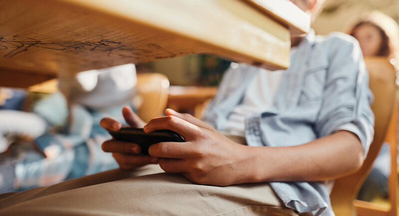 A teenage student sitting at a desk, discreetly holding their cell phone underneath.