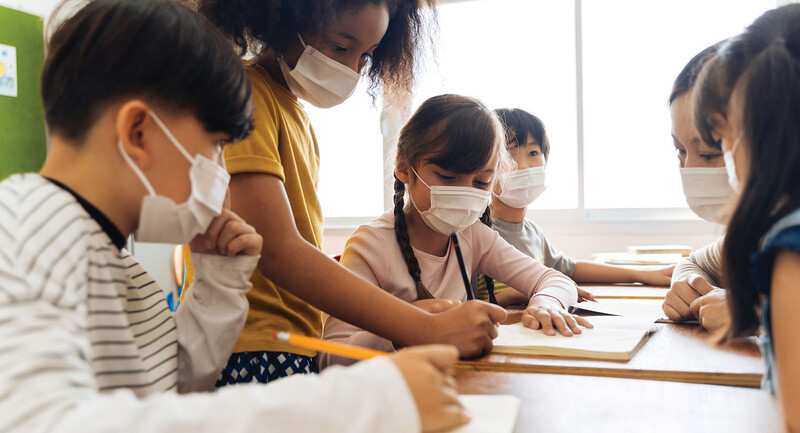 Photo of young students gathered around a table wearing masks