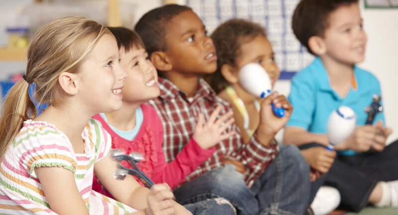 A group of children sits on the floor in a classroom smiling happily and playing musical instruments