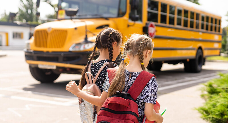 Two children talk to each other and walk to school with their backs turned toward the camera, a school bus in the background behind them